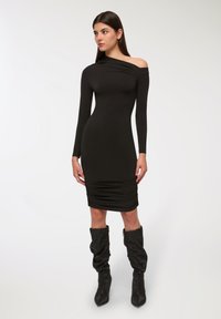 Abito nero aderente con maniche lunghe e scollatura asimmetrica. Tessuto a coste, dettagli arricciati all'orlo, abbinato a stivali al ginocchio.