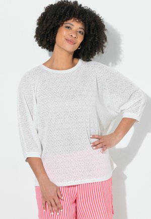 Vrouw met krullend haar, gekleed in een wit getextureerd topje met driekwart mouwen en rood-wit gestreepte broek, poseert met één hand op haar heup.