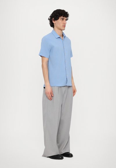 Camicia azzurro chiaro a maniche corte con bottoni, abbinata a pantaloni grigi a gamba larga e scarpe nere basse. Design semplice e dalla vestibilità rilassata.