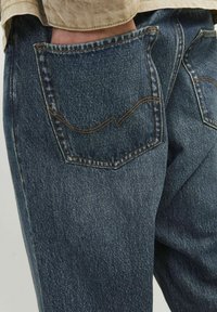 Los jeans de mezclilla azul oscuro presentan bolsillos traseros con costuras de contraste, un ajuste relajado y una textura ligeramente desgastada.