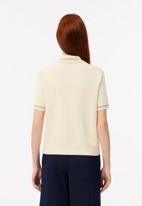 Polo shirt crema a maniche corte con texture a coste, caratterizzato da un colletto stretto con strisce verdi e gialle, e un taglio aderente.