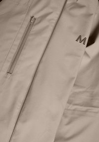 Abrigo beige con un bolsillo con cremallera y un logo abstracto. Textura suave, diseño elegante con una forma ajustada y acentos minimalistas.