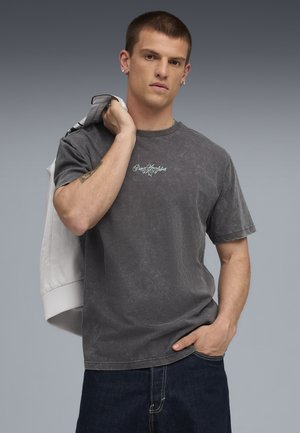 Ung mand iført en falmet sort Puma Sportstyle t-shirt, mørke jeans, som holder en hvid jakke over sin skulder mod en grå baggrund.