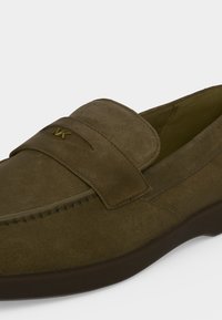 Mocassin en daim marron avec un bout arrondi, des détails cousus, une bride sur le dessus et un petit logo MK doré sur la bride.