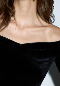 Top negro de hombros descubiertos hecho de un tejido suave y brillante. Presenta un escote amplio y un diseño ajustado que resalta la zona de las clavículas.