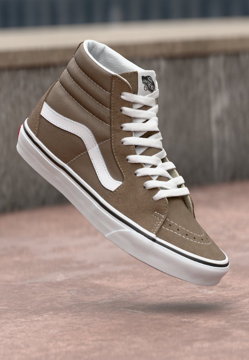 High-top sneakers in brown suede with white side stripe, textured paneling, white laces, and a rubber sole with a red accent on the heel.
