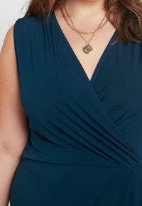 Marinblå omslagsklänning med korta ärmar, med samlad struktur vid midjan och subtil detaljering vid axlarna. Inkluderad guldhalsband i lager.