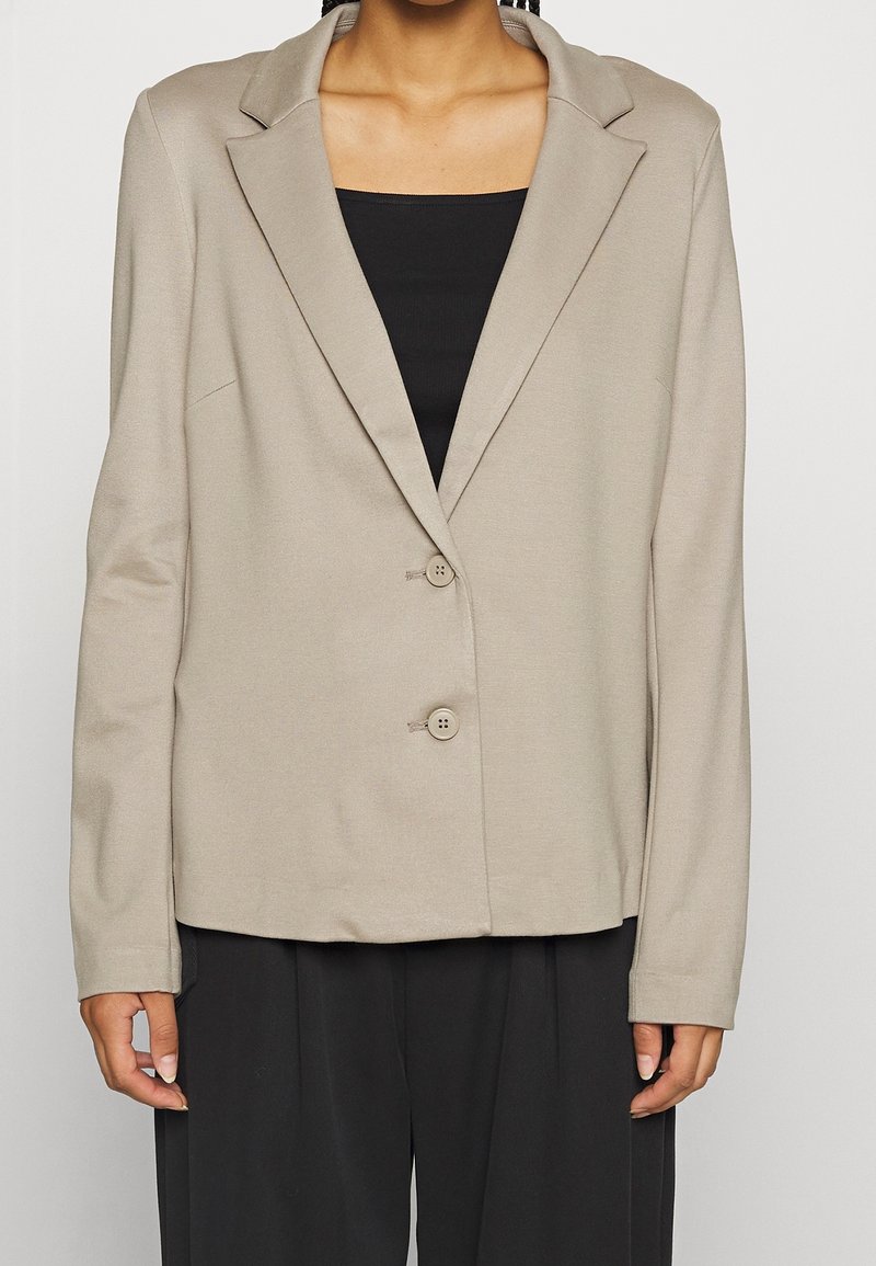 Vue de face d'une personne portant un blazer beige à deux boutons sur un haut noir et un pantalon noir, les mains détendues le long du corps.