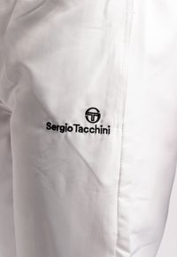 Bijele atletske hlače s teksturiranim materijalom, s crnim izvezenim logotipom "Sergio Tacchini" sa strane. Jednostavan i elegantan dizajn.