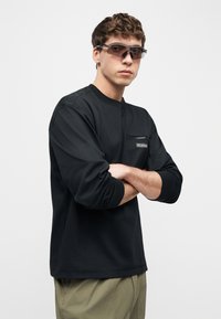 Νεαρός άνδρας που φοράει μαύρο μακρυμάνικο πουκάμισο με τσέπη στο στήθος με φερμουάρ και αθλητικά γυαλιά ηλίου με καφέ απόχρωση, με σταυρωμένα χέρια, στέκεται μπροστά σε απλό λευκό φόντο.