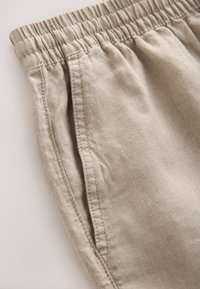 Pantaloni in lino beige con vita elasticizzata, dotati di due tasche laterali e cuciture visibili. Il tessuto ha una finitura morbida e testurizzata.