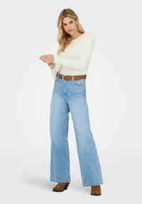 Blonde vrouw in een witte langemouwen top, hoge taille wijde lichtblauwe jeans, bruine riem en bruine haklaarzen, staande tegen een witte achtergrond.