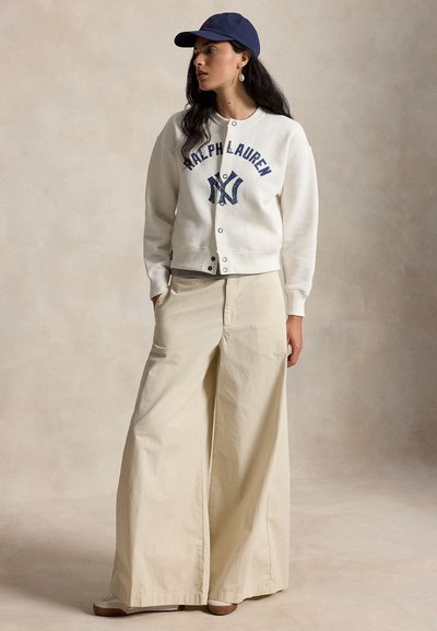 Γυναίκα που φοράει ένα λευκό φούτερ Ralph Lauren, μπεζ παντελόνια με φαρδιά πόδια, μπεζ παπούτσια και μπλε σκούφο μπέιζμπολ, κοιτάζοντας προς το πλάι.