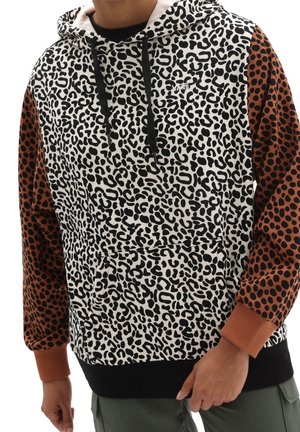 Mand iført en hættetrøje med hvidt og sort leopardmønster foran og brune ærmer med sorte pletter, hænder afslappet ved siderne.