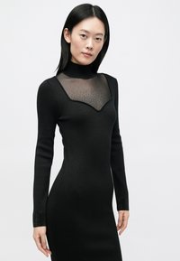 Robe noire moulante avec un col montant, un panneau transparent au niveau de la poitrine orné de petits pois, et des poignets texturés embellis de sequins.