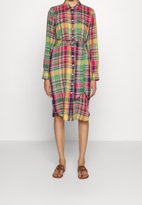 Eine Frau trägt ein kariertes, mehrfarbiges Hemdkleid mit langen Ärmeln, einem Taillengürtel und braunen Sandalen mit offenen Zehen und steht vor einem schlichten Hintergrund.