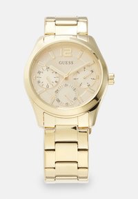 Guldfarvet armbåndsur med en tekstureret rund urskive, markante timeindikatorer, tre undermålere og et metal-link armbånd. Mærket "Guess" vises.