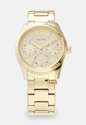 Goldfarbene Armbanduhr mit einem strukturierten runden Zifferblatt, auffälligen Stundenmarkierungen, drei Teilzifferblättern und einem Metallgliederarmband. Markenname "Guess" angezeigt.