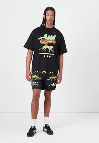 Melnā oversize T-kreklā ir koša grafiskais dizains ar leopardu un tekstu. Melnās šortos ir leopardu raksts un logo detaļa, saskaņoti ar melnajām sporta kurpēm.