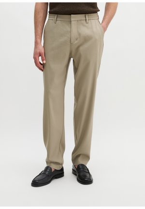 Man draagt beige broek, zwarte loafers en een donkerbruin gebreide top, staand met één hand in de zak.