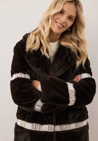 Donkerbruin nepbont jas met een brede kraag, oversized mouwen en witte merkaccenten op de riem en manchetten. Zachte textuur.