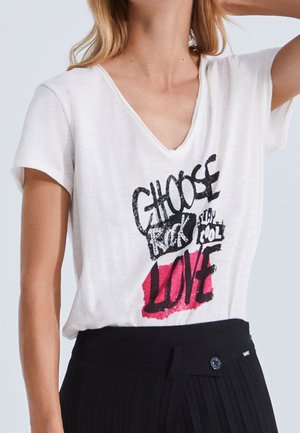 Kobieta ubrana w białą koszulkę z dekoltem w serek z czarnym i czerwonym napisem "Choose Rock Stay Cool Love", włożoną do wysokich, czarnych plisowanych spodni.