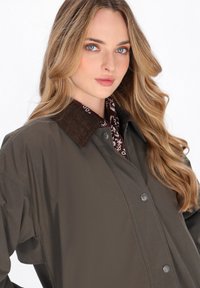 Donkerbruine jas met een corduroy kraag, knopen aan de voorkant en een patroon-sjaal zichtbaar eronder. Glad, gestructureerd materiaal.