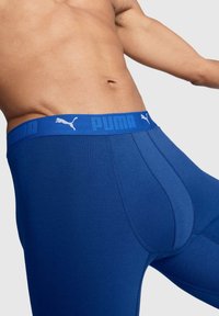 Közelkép egy férfi törzséről, aki kék Puma boxer alsót visel, logózott derékpánttal egy sima szürke háttér előtt.
