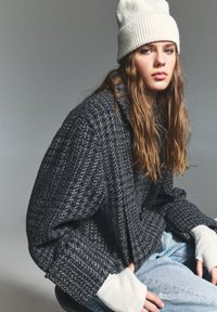Cardigan tricotat gri texturat, cu croială supradimensionată, având mansete ribbate. Asortat cu o căciulă tricotată gri deschis și blugi uzati de culoare albastră.