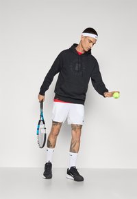 Black hoodie with logo, red shirt underneath, white shorts, black sneakers, white socks, holding a tennis racket and tennis ball.