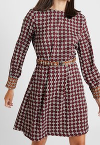 Une robe moulante bordeaux à manches longues avec des motifs géométriques blancs et une ceinture orange. Les manches ont des poignets avec un motif contrastant.