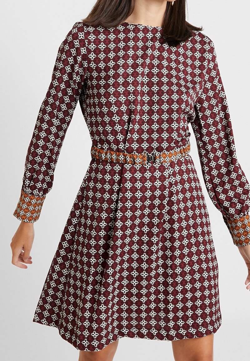 Une robe moulante bordeaux à manches longues avec des motifs géométriques blancs et une ceinture orange. Les manches ont des poignets avec un motif contrastant.