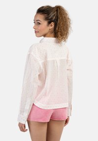 Camicia bianca con bottoni, decorata con pois colorati, maniche lunghe e dettagli a yoke. Indossata con pantaloni corti rosa, che mostrano una vestibilità comoda.