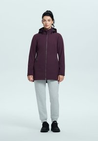 Giacca impermeabile viola con zip frontale, cappuccio e due tasche laterali, abbinata a pantaloni felpati grigi e scarpe da ginnastica nere.