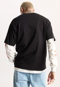 Camiseta negra de cuello redondo superpuesta sobre una camisa blanca de manga larga con acentos gráficos en marrón claro. Jeans de mezclilla, con bolsillos traseros visibles.