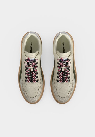 Paire de baskets beige avec bouts texturés, lacets multicolores noirs, blancs et rouges, semelles couleur marron clair et nom de la marque imprimé à l’intérieur.