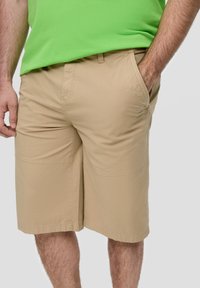 Shorts cargo beiges avec une texture lisse, dotés de poches latérales et d'une coupe décontractée. Portés avec une chemise verte, mettant en avant un style décontracté.