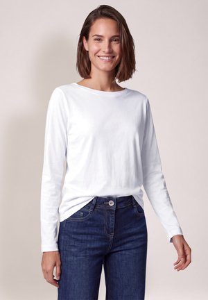 Mujer sonriente con una camisa blanca lisa de manga larga y vaqueros azul oscuro, de pie frente a un fondo claro neutro.