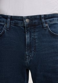 Jeans in denim blu scuro con una texture liscia, dotati di chiusura a bottone, passanti per cintura e dettagli sulle tasche frontali.