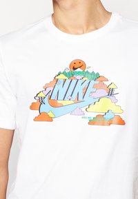 Camiseta blanca de algodón con un colorido logo de Nike que incluye nubes, montañas y un gráfico de un sol sonriente, mostrando tonos vibrantes y un diseño divertido.