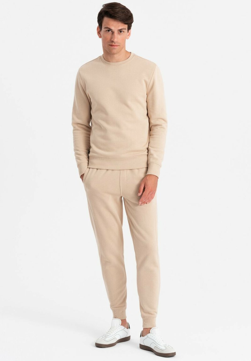 Beiges Loungewear-Set bestehend aus einem gerippten, langärmeligen Rundhals-Sweatshirt und passenden tapered Hosen, kombiniert mit weißen Sneakers.