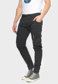 Zwarte cargobroek van katoenblend, met zijzakken, ritsdetails op het been, slim fit en elastische manchetten. Gepaard met grijze sneakers.