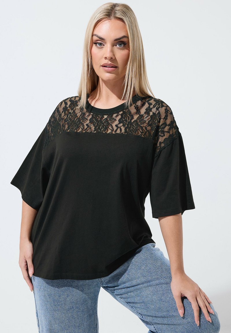 Mujer rubia con blusa negra con canesú de encaje floral y mangas sueltas, combinada con jeans azul claro, posando frente a un fondo liso.