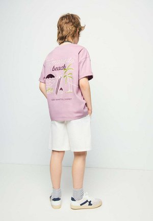 Ragazzo con capelli castano chiaro sta con le mani nelle tasche, indossando una maglietta lilla a tema spiaggia, pantaloncini bianchi, calzini grigi e scarpe da ginnastica bianche.