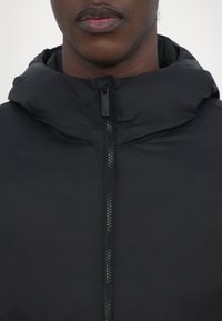 Schwarze gepolsterte Jacke mit Reißverschluss und Kapuze. Hat eine weiche Textur und ein minimales Design, geeignet für kaltes Wetter.