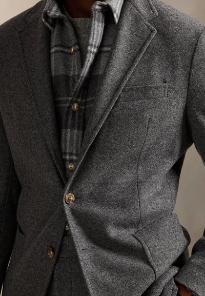 Man draagt een gelaagde grijze wollen blazer over een grijs geruit overhemd met knopen en een trui, poserend met één hand in de zak.