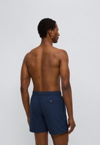 Μακό σορτς κολύμβησης σε ναυτικό μπλε με ελαστική μέση, διαθέτει μικρή πίσω τσέπη με τρίχρωμη λεπτομέρεια. Υφασμένο ύφασμα με κάθετες ρίγες.