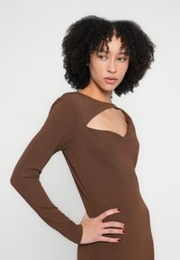 Brūna, garām piedurknēm kleita ar izgriezuma kakla izgriezumu. Gluda tekstūra, pieskaņota pie ķermeņa un minimālistisks dizains, stilizēta ar roku uz gurna.