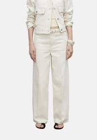 White denim jacket with two chest pockets, paired with loose-fitting white pants. Features a straight cut and worn with black sandals.