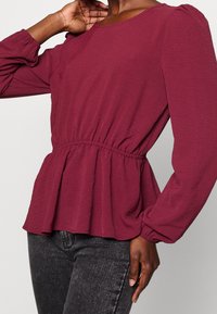 Kvinna klädd i en långärmad vinröd peplumtop med gathered midja och svarta tvättade jeans, poserande med en hand nära sin hals.